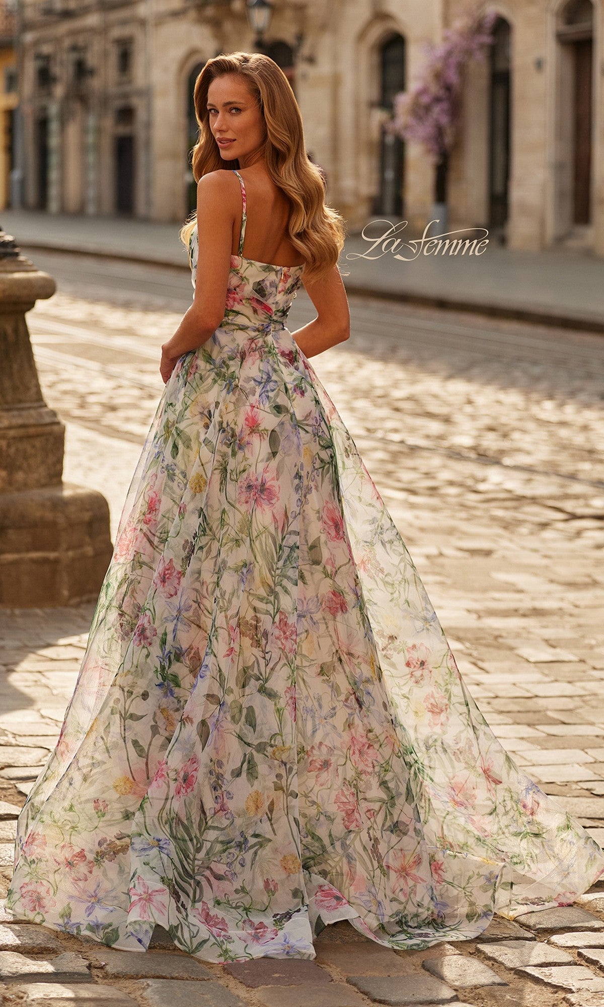 A woman stands outdoors on a cobblestone street, looking over her shoulder in the La Femme 33516 long prom dress with a flowing floral a-line skirt. Historic stone buildings are visible in the background.